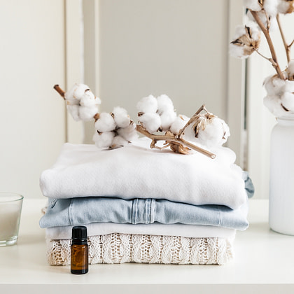 a stack of cotton clothes, a bottle of essential oil and a vase of cotton flowers