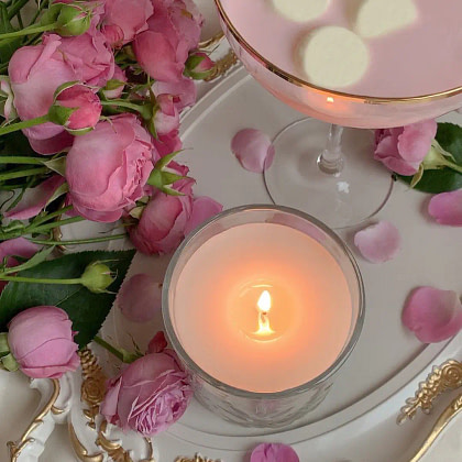 A pink cocktail alongside a candle is displayed on a tray, complemented by delicate pink roses.